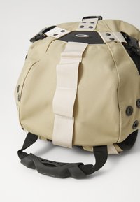 Beige Stofftasche mit schwarzen Akzenten, ausgestattet mit einer strukturierten Oberfläche, verstellbaren Trägern und einem gummierten Griff für besseren Halt.