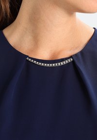 Marinskräddad krage med en rad små, klara rhinestones längs halsringningen. Mjuk tyget med en slät yta och elegant fall.