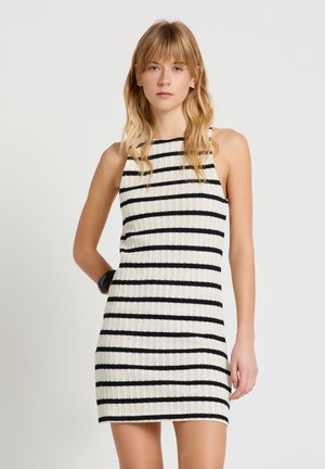 Donna con capelli biondi fino alle spalle che indossa un vestito bianco senza maniche in maglia con strisce nere orizzontali su uno sfondo chiaro e uniforme.