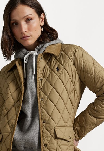 Beige gewatteerde jas met een corduroy kraag en drukknoopsluiting, gedragen over een grijze hoodie. Heeft een klein logo op de borst en een gemêleerde stof.