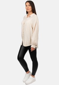 Camicia beige con bottoni, in tessuto testurizzato e con motivi bianchi, abbinata a leggings neri e sneakers bianche, che evidenziano un fit rilassato.