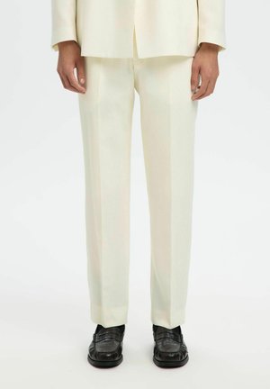 Dolna połowa osoby ubranej w kremowe, dopasowane spodnie z pasującą marynarką oraz czarne skórzane mokasyny na jednolitym tle.