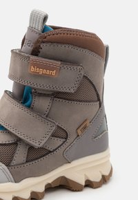 Szürke bőr és textil gyermekcipő tépőzáras pántokkal, barna kiegészítőkkel és texturált talppal, amelynek kontrasztos tejfehér alapja van.