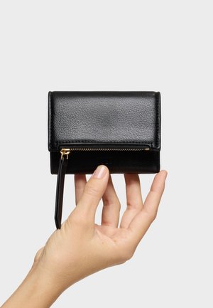 Mano sosteniendo una pequeña cartera de cuero negra con una cremallera y tirador dorados sobre un fondo liso.