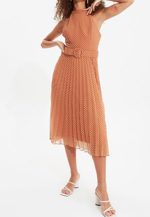 Brązowa sukienka w groszki z plisowanym dołem, dekoltem halter i paskiem. Materiał jest lekki i półprzezroczysty. W połączeniu z białymi sandałami.