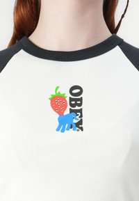 Valge soonikkoes T-särk mustade raglanvarrukatega. Omab värvilist graafikat maasikast ja sinisest elevandist, koos "OBEY" tekstiga.