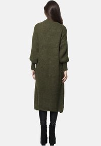 Lange, olivgrüne Strickjacke, mit strukturiertem Muster, übergroßen Ärmeln und seitlichen Schlitzen, getragen über schwarzen, eng anliegenden Hosen und Stiefeletten.