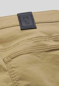 Khakifarbenes Baumwollhosen mit einem navyblauen Lederlabel mit "M5", kontrastierenden Nähten und seitlichen Taschen auf einer glatten Textur.