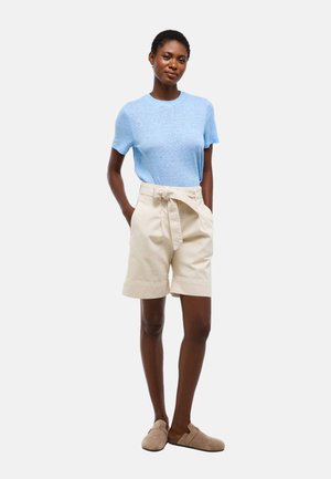 Femme debout portant une chemise bleu clair à manches courtes, un short beige taille haute avec ceinture, et des chaussures beiges à enfiler sur fond blanc.