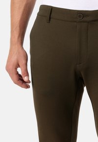 Pantaloni verde oliva con una texture liscia, dotati di chiusura con bottone frontale e tasche laterali. Vista parziale di una mano sul tessuto.
