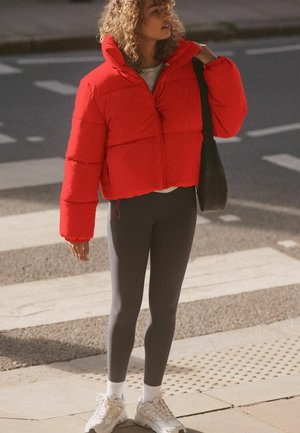 Červená skrátená puffa bunda s vysokým golierom a elastickými manžetami, v kombinácii so sivými legínami a bielymi teniskami. Na pleci jednoduchá čierna taška.