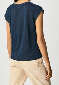 Námorně modrá košile s krátkým rukávem a uvolněným střihem, s kulatým výstřihem, vyrobená z lehkého materiálu, zobrazená zezadu s světlými kalhotami.