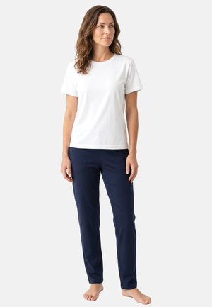 Woman standing barefoot wearing a plain white short-sleeve T-shirt and dark blue straight-leg pants against a neutral background.