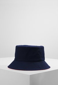 Cappello a secchiello blu navy realizzato in tessuto morbido, con una visiera ampia e inclinata verso il basso, e dettagli rossi lungo il bordo. Finitura in tessuto texture.