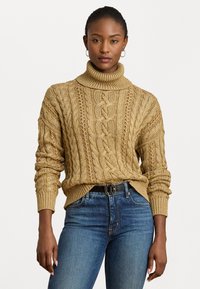 Vītā turtlenecks džemperis zeltaini bēšā tonī, ar teksturētu rakstu, ribotām piedurknēm un apakšmalu, apvienots ar ziliem džinsiem.