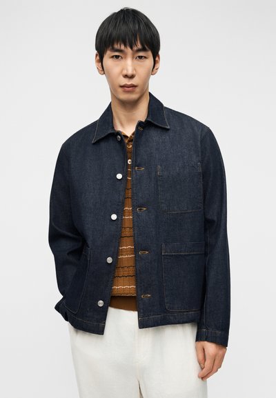 Άνδρας που φοράει σκούρο τζιν μπουφάν πάνω από καφέ πουκάμισο με σχέδια και λευκό παντελόνι, στέκεται μπροστά σε απλό φωτεινό φόντο.