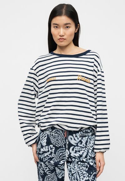 Ριγέ πουκάμισο με μακριά μανίκια σε λευκό και ναυτικό μπλε, με κεντημένο λογότυπο "Rebels" σε πορτοκαλί. Συνδυάζεται με τζιν παντελόνι που έχει λευκά μοτίβα κειμένου.