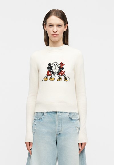 Femme portant un pull blanc avec un motif Mickey et Minnie Mouse en train de s'embrasser, associé à un jean large bleu clair avec une légère usure.