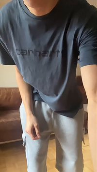 Άτομο που φοράει ένα σκουρόχρωμο t-shirt Carhartt και ανοιχτό γκρι παντελόνι, στέκεται μέσα σε κλειστό χώρο κοντά σε ένα καφέ δερμάτινο καναπέ πάνω σε ξύλινο πάτωμα.
