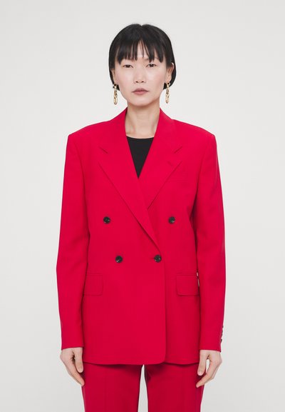 Blazer rouge à double boutonnage avec revers en pointe, poches latérales et boutons noirs. Tissu lisse avec une coupe ajustée, associé à un haut noir.