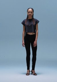 Grijze cropped shirt, zwarte skinny jeans en zwarte hoge hakken. Model staat tegen een lichtblauwe achtergrond. Haar in vlechten.