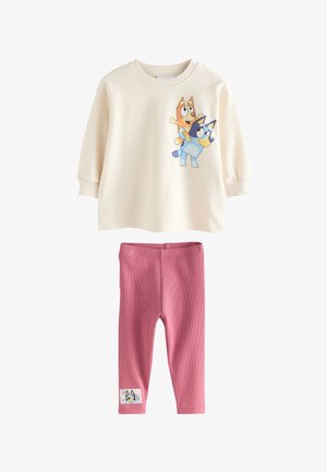 Barn med blondt hår i en lav knold iført en cremefarvet sweatshirt med et farverigt Bluey tegneseriehund-design på ryggen.