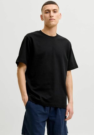 Ung mann med kort hår iført en ensfarget svart t-skjorte og marineblå shorts, stående med den ene hånden i lomma mot en nøytral bakgrunn.