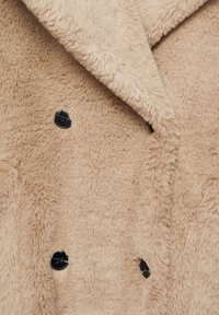 Manteau en fausse fourrure beige avec une texture moelleuse, présentant un design croisé et des boutons noirs, exprimant chaleur et douceur.