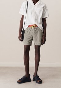 Camisa blanca de manga corta con tela texturizada, pantalones cortos grises con cordón amarillo, y sandalias negras; accesorios mínimos, diseño sencillo.