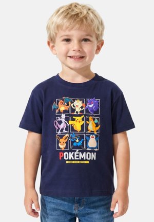 Jeune garçon souriant portant un t-shirt bleu marine avec des personnages colorés de Pokémon et le texte « Pokémon prêt pour le combat ».