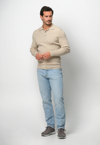 Beige Langarm-Polohemd mit Kragen und drei Knöpfen, kombiniert mit hellblauen Jeans und grauen Sportschuhen, vor einem weißen Hintergrund stehend.