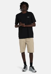 Joven de pie, vistiendo una camiseta negra con texto, pantalones cortos beige, calcetas negras y zapatillas plateadas, contra un fondo blanco liso.