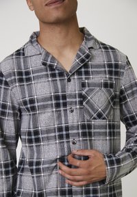Man in een grijs-zwarte geruite overhemden met knoopsluiting en een borstzak, die het shirt met één hand bij de knopen vasthoudt.