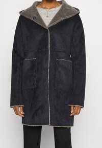 Cappotto blu navy scuro con una morbida fodera in pelliccia grigia e ampie tasche frontali. Presenta un cappuccio alla moda e dettagli di cucitura in evidenza.