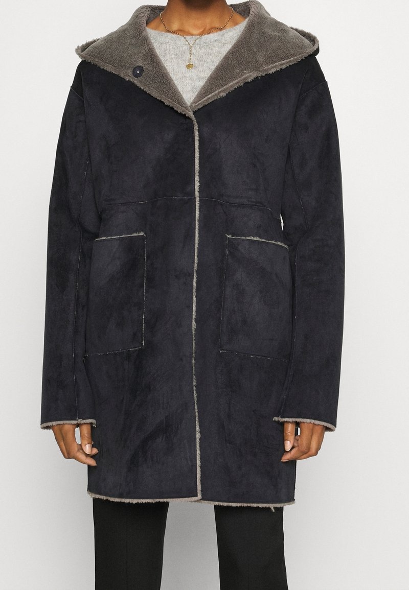 Cappotto blu navy scuro con una morbida fodera in pelliccia grigia e ampie tasche frontali. Presenta un cappuccio alla moda e dettagli di cucitura in evidenza.