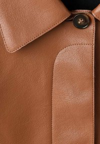Detalle de chaqueta de cuero marrón que muestra un gran cuello, superficie texturizada y un cierre de botón clásico, destacando la costura y el diseño de paneles.