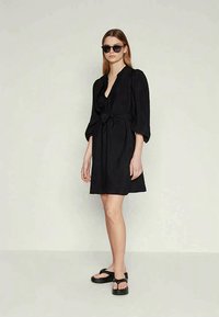 Juoda suknelė su gilia V formos iškirpte, diržu ant liemens ir pūstomis rankovėmis. Stilinga su juodais saulės akinių akiniais ir masyviomis juodomis šlepetėmis. Lygi tekstūra.