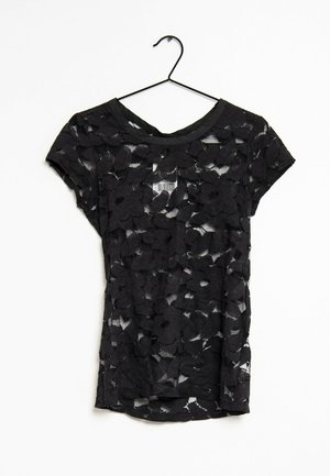 Haut noir à manches courtes avec motif en dentelle florale, tissu transparent, encolure ronde et ourlet arrondi, suspendu à un crochet noir.