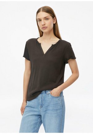 Femme aux cheveux longs et raides portant un T-shirt noir à manches courtes avec un col en V et un jean bleu clair, debout avec une main dans la poche devant un fond blanc.