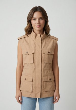 Femme portant un gilet utilitaire beige sans manches avec des poches à rabat boutonnées sur un jean bleu clair, se tenant devant un fond uni.