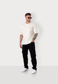 Cream cotton t-shirt with short sleeves and logo, paired with black cargo pants featuring side pockets, worn with white sneakers and sunglasses.