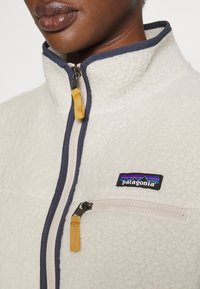 Fleece jakna u krem boji s mornarskom plavom obrubom, koja sadrži džep sa zipom i žutom ručkom te Patagonia logotipom na prsima.