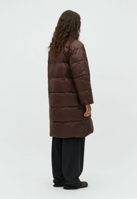 Cappotto imbottito marrone con un design lungo e quadrato, caratterizzato da sezioni trapuntate. Il tessuto appare liscio e lucido, con maniche lunghe e tasche.