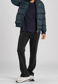 Mörkgrön vadderad jacka med dragkedja, svarta byxor och ljusa sneakers. Jackan har texturerad detalj och en logotyp på övre vänstra sidan.