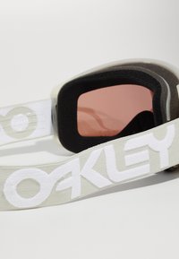 Beskyttelsesbriller med svart skuminnside, rosa linse og bred, lysegrå stropp med den hvite "OAKLEY" logoen. Glatt, fleksibel design.