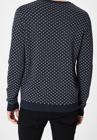 Marinblå tröja med små vita snöflinge-mönster, som bärs av en person som vänder ryggen mot kameran, i kombination med svarta byxor.