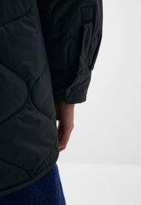 Svart quiltad jacka med en slät textur, sned ärmsöm och sidofickdetalj, som bärs över blå denim.