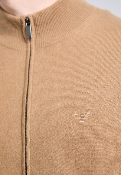 Tuvumā redzama bēša krāsā zip-up džemperis ar ribotu apkakli un mazu izšūtu logotipu uz krūtīm, ko valkā cilvēks.