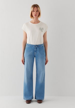 Woman with short blonde hair wearing a white t-shirt with a small chest design and light blue wide-leg jeans standing against a plain white wall.