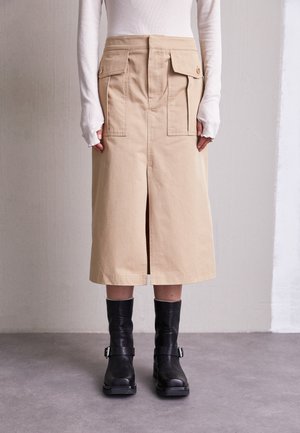 Beżowa spódnica do kolan z przednim rozcięciem i kieszeniami na klapki, zestawiona z białą bluzką z długim rękawem oraz czarnymi, sięgającymi połowy łydki butami na klamry.